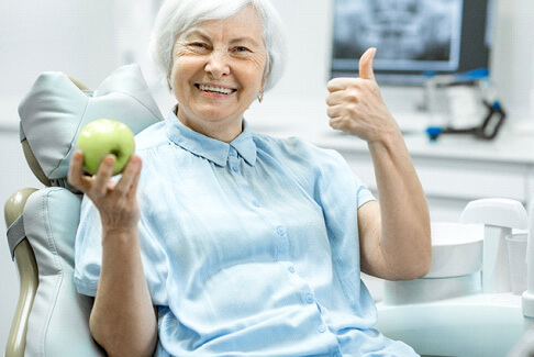woman in pale blue shirt in dental chair holding green apple giving thumbs up