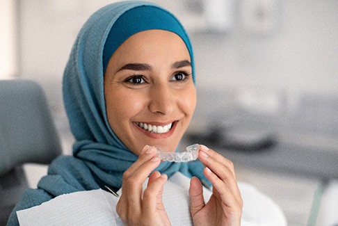 Woman in blue head cover sitting in dental chair smiling holding Invisalign